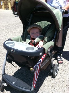6 month old white baby boy in large green stroller with canopy and tray. Small American flag sticking out of stroller.